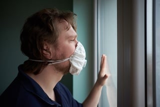 Caucasian man wearing protective medical mask looking out the window during quarantine