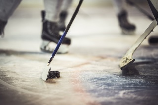 Close up of hockey puck and stick during a match with unrecognizable players in the background.