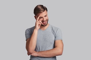 Good looking young man smiling and touching his head with hand while standing against grey background