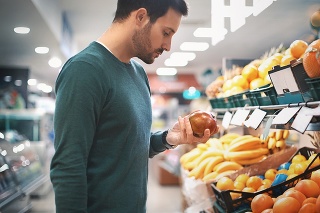 Closeup side view of a handsome early 30's man buying some fruit at a local supermarket. He's holding a pomegranate and thinking if it's ripe. Large stack of oranges, bananas and pineapples in front of him.