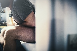 Closeup of mid 30's unrecognizable man having stomach cramps. He's in toilet, holding his belly and making some painful grimaces. Partially obscured by bathroom door, toned image. Side view.