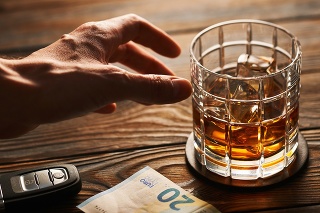 Man's hand reaching to glass of whiskey or alcohol drink with ice cubes and car key on rustic wooden table. Drink and drive and alcoholism concept. Safe and responsible driving concept.