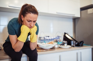 Young housewife is sitting on the kitchen counter, wearing protective gloves, tired from washing dishes.