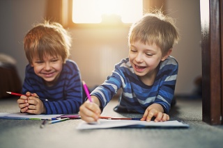 Two happy little boys aged 5 drawing on the floor. Sun is backlighting the kids from the window behind them with warm light.