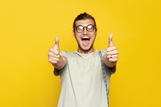 Handsome cheerful man with eyeglasses smiling and showing thumb up gesture with both hands while standing on yellow background and looking at camera.