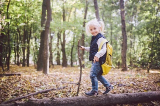 Theme outdoor activities in nature. Funny little Caucasian blonde girl walks walks hiking in the forest on rough terrain with a large backpack. Uses walking stick.