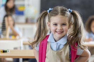 Pretty Caucasian preschool age girl stands in her classroom on the first day of school. She is wearing a school uniform and has blond hair in ponytails.