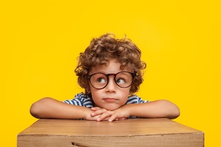 Portrait of curly boy in glasses leaning on box looking away pensively on orange background.