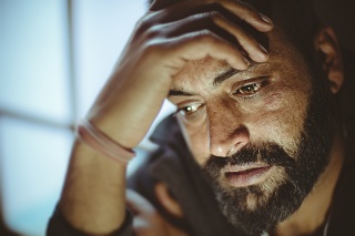 Indoor close-up image of disturbed, sad, Asian, Indian mid adult man with strong character and facial hair. He is sitting at home near door in day time. He is crying and a drop of tear coming out of his eye. He is looking down and holding his head while thinking something deeply with blank expression.