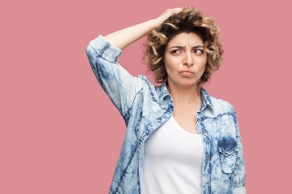Portrait of thoughtful confused young woman with curly hairstyle in casual blue shirt standing scratching her head and thinking what to do. indoor studio shot, isolated on pink background.