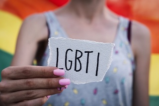closeup of a young caucasian woman holding a paper note in front of her with the text LGBTI written in it, with a rainbow flag in the background