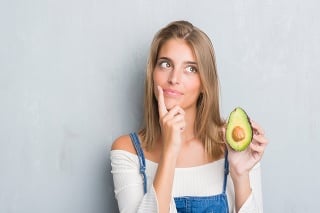 Beautiful young woman over grunge grey wall eating avocado serious face thinking about question, very confused idea