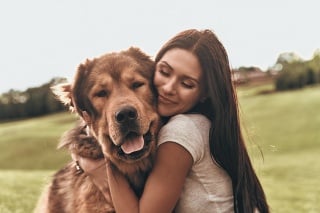 Beautiful young woman keeping eyes closed and smiling while embracing her dog outdoors
