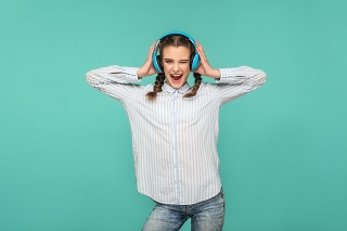 Happy funny girl in striped blue shirt and pigtail hairstyle, standing listening music with headphone, looking at camera with safisfied face, Indoor studio shot, isolated on blue or green background