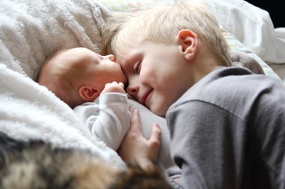 A 5 year old big brother is hugging, smiling, and looking at his newborn baby sister as they sunggle in bed.