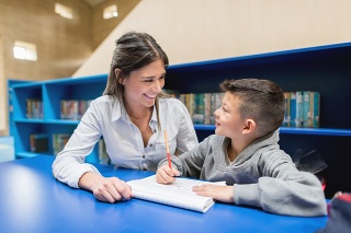 Happy teacher with a little student at the library doing his homework - education concepts