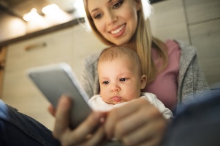 Beautiful young blond mother at home with her little baby son in the arms, sitting on the kitchen floor, holding smart phone, smiling