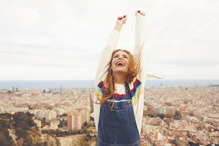 Happy woman with arms raised looking up. Young female is standing against cityscape. She is wearing casuals.