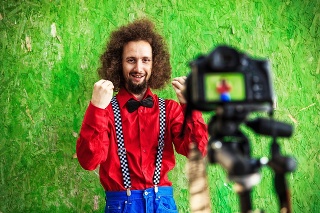 Young man standing in front of green background and vlogging. Wears red shirt and blue  trousers, bowtie and suspenders.