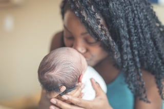 Close-up of an African American mom as she gently cradles and kisses her newborn daughter in her arms. She is wrapped up and swaddled and is sleeping with a content expression. Mom has her hands interlocked behind baby's full head of hair as she snuggles with her.