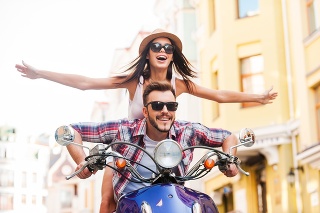 Beautiful young couple riding scooter together while happy woman keeping arms outstretched and smiling