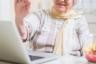 Gray-haired senior woman waving hand in front of laptop while having video call with her family members.