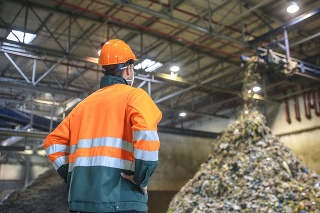 Low angle rear view of young male worker in helmet, pollution mask, and reflective clothing observing waste falling from conveyor belt onto pile at facility.