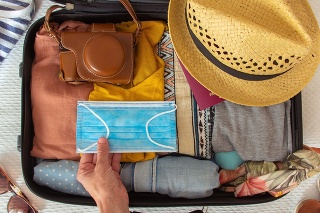 Hand putting a face mask on the suitcase prepared to travel in summer, in the new normal, after the coronavirus covid 19 pandemic. There are other accesories like a hat, a camera and a bikini. Concept of quarantine, coronavirus, summer vacation in new normal. Flat lay or top view
