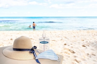 Vacationing in the New Normal after COVID-19 coronavirus pandemic. Tourism concept showing lounge chair on sandy beach with beach hat, hand sanitizer, medical face mask and copy space.