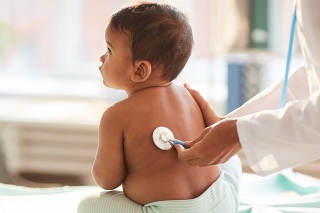 Rear view of shirtless toddler sitting on the table while doctor listening to him with stethoscope