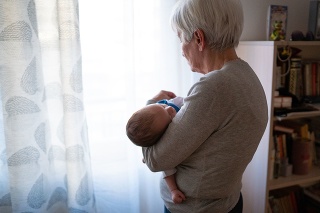 Rear view of grandmother holding newborn grandson in her arms while standing and looking through the window at home