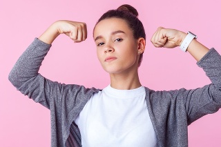 Portrait of self-confident teenage girl with bun hairstyle in casual clothes raising hands showing power, feeling independent strong with proud look. indoor studio shot isolated on pink background