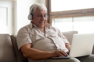 Smiling older man wearing headphones using laptop at home, looking at screen, listening to favorite music and searching information in internet, happy mature male making video call, sitting on couch