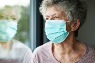 a old woman or grandma is wearing a respirator or surgical mask and is looking out the window while she is in quarantine because of the corona virus