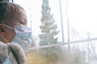 Sad illness child on home quarantine. Boy and his teddy bear both in protective medical masks sits on windowsill and looks out window. Virus protection, coronavirus pandemic, prevention epidemic.