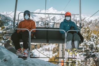 two skiing people, man and woman wearing mouth nose mask on chair ski lift with social distance on sunny winter day in the mountains