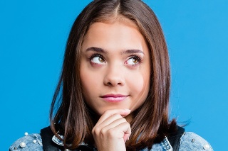 Cute female high school student wearing oversized denim jacket and white t-shirt standing against blue background. Portrait of pensive teenager with hand on chin.