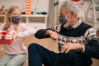 Little blonde girl receiving a Christmas present from her grandfather, both wearing a protective face mask, maintaining social distancing and thanking him for the present by bumping an elbow with him