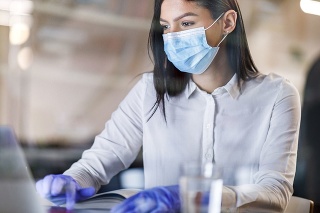 Female entrepreneur with face mask and protective gloves working on a computer during corona virus pandemic in the office. The view is through glass.