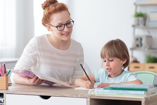 Smiling mother helping son with exercises of math while sitting at desk with notebooks