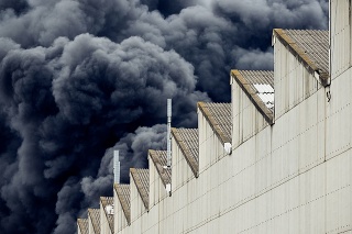 A toxic fire in an industrial district creates plumes of dangerous smoke billowing up into the air as seen from behind a near by warehouse building.