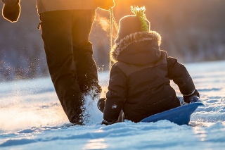 A grandfather is pulling his grandson on a sled on a frozen lake in winter at sunset. They are both having fun. There is a mountain range in the background.