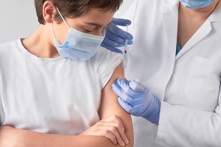 Corona virus, flu or measles vaccine concept. Medic, doctor, nurse, health practitioner vaccinates teenage boy with vaccine in syringe. She is wearing uniform, disposable hut, blue gloves and face mask