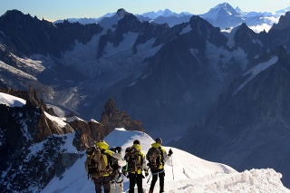 DSLR picture of four Alpine climbers going down the ridge at l'Aiguille du Midi to reach the Mont Blanc summit. The climbers are on the foreground and the alps are in the background and the sky is clear.