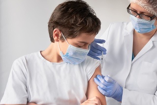 Corona virus, flu or measles vaccine concept. Medic, doctor, nurse, health practitioner vaccinates teenage boy with vaccine in syringe. She is wearing uniform, disposable hut, blue gloves and face mask