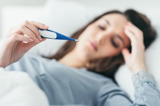 Woman with flu virus lying in bed, she is measuring her temperature with a thermometer and touching her forehead