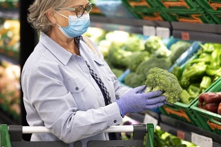 Senior woman wearing face mask and rubber gloves selects broccoli and other vegetables  in a supermarket -active elderly pensioners with trolley