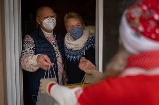 the new normal in times of corona virus crisis with love and positive emotions, celebrating christmas with social distance, happy smiling behind the mask senior couple, parents  giving christmas presents to their daughter blurred in foreground at open door of their house