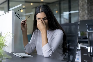 Portrait of depressed young Latin-American businesswoman sitting at computer in office holding glasses and rubbing eyes. Overworking concept