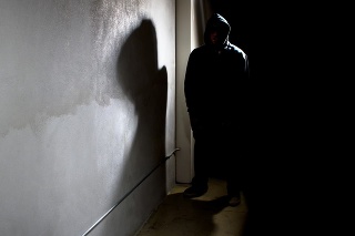 Photo of a hooded criminal stalking in the shadows of a dark street alley.  The hooded man is a silhouette and hiding in the dark.  The man is a criminal waiting to ambush victims.  The concrete walls provide copyspace.The photo depicts crime.